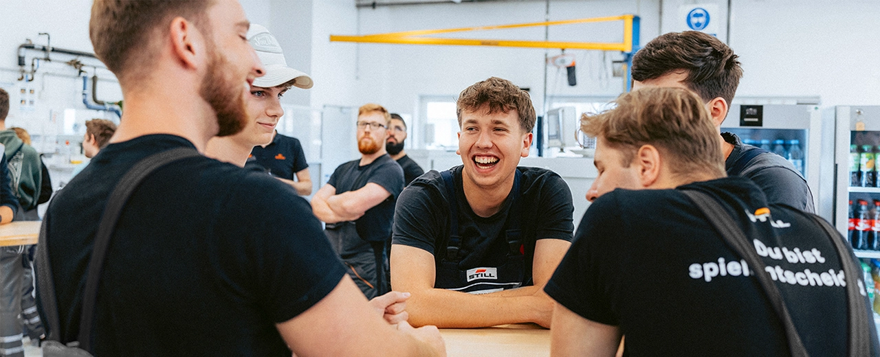 Eine Gruppe junger STILL-Mitarbeiter in schwarzer Arbeitskleidung unterhält sich lachend an einem Tisch in einer Werkstatt.