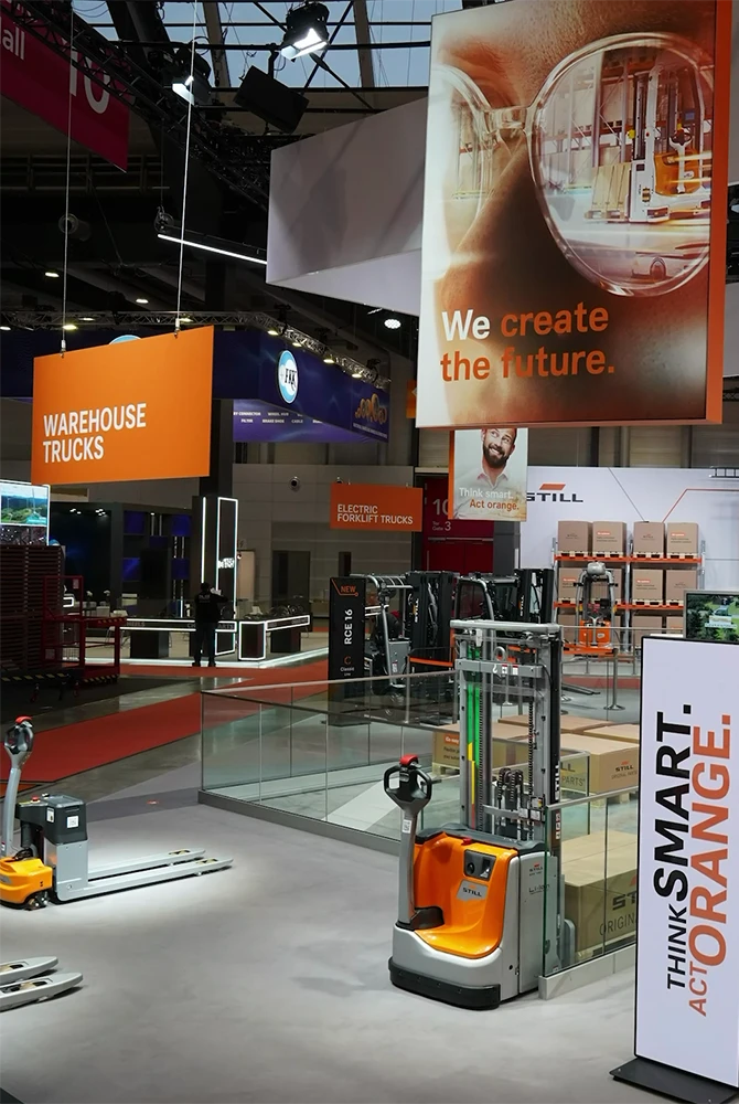 Messestand von STILL mit orange-grauen Flurförderzeugen, einem Schild „Warehouse Trucks“ und Bannern mit den Slogans „We create the future.“ und „Think smart. Act orange.“