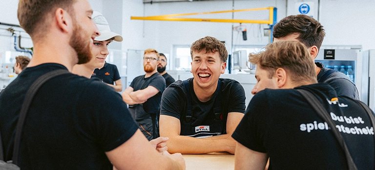 A group of young STILL employees in black work clothes chat and laugh around a table in a workshop.