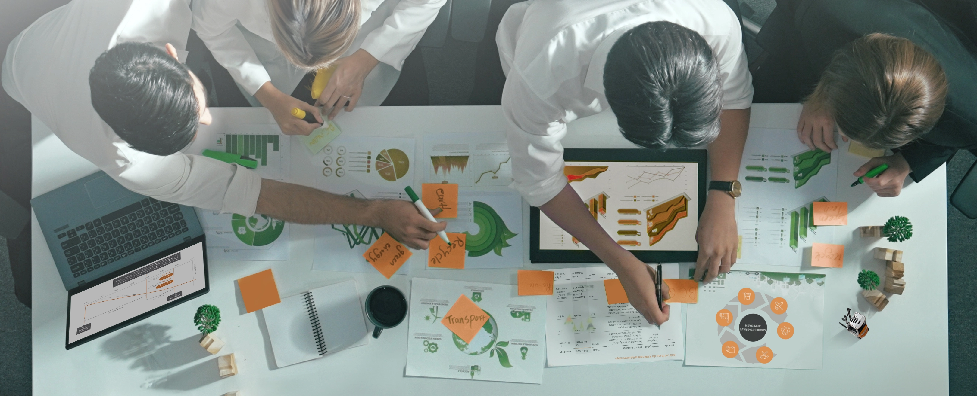 Top view of four people working at a table covered with sustainability charts, sticky notes and a laptop.