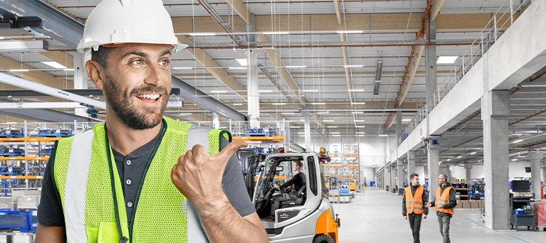 A smiling man in a hard hat and high-visibility vest points with his thumb towards a warehouse, where a grey and orange STILL forklift is working and two colleagues in safety vests are walking.