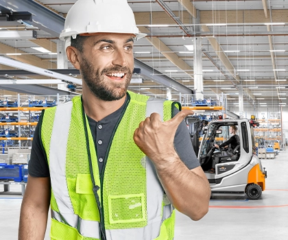 A smiling man in a hard hat and high-visibility vest points with his thumb towards a warehouse, where a grey and orange STILL forklift is working and two colleagues in safety vests are walking.