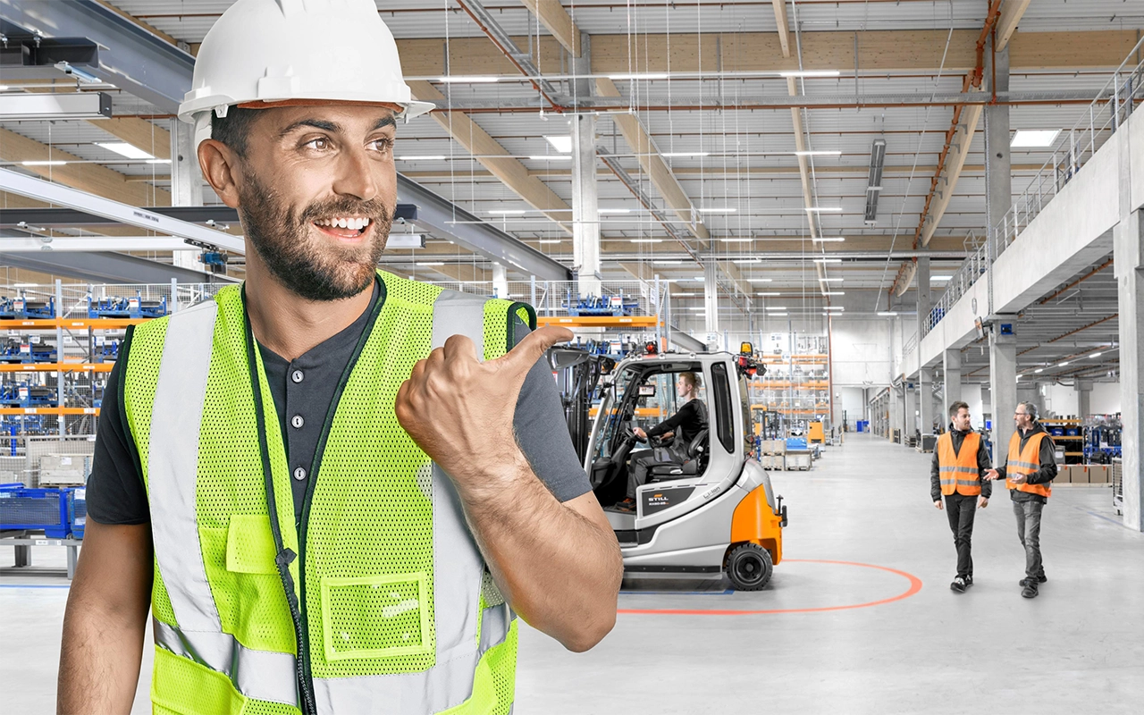 A smiling man in a hard hat and high-visibility vest points with his thumb towards a warehouse, where a grey and orange STILL forklift is working and two colleagues in safety vests are walking.