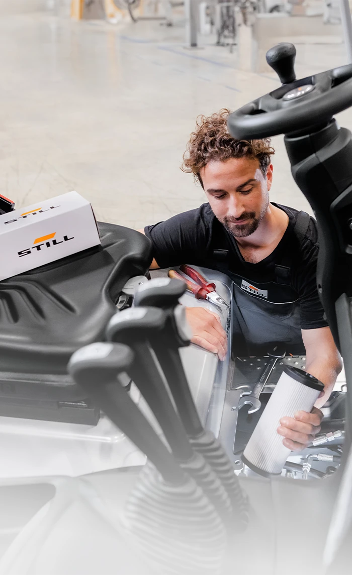A STILL service technician holds a filter cartridge while servicing a forklift, with a STILL spare parts box on the driver’s seat.
