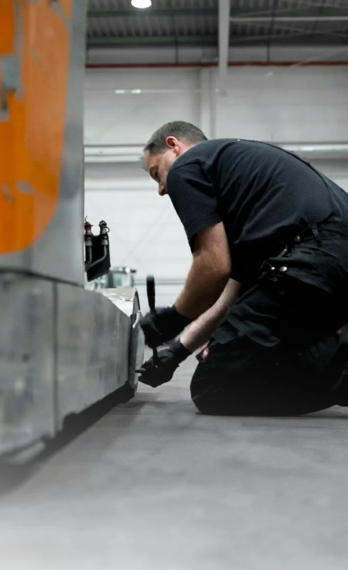 A service technician in black workwear kneels on the floor of a warehouse and works on an orange and grey industrial truck with a tool.