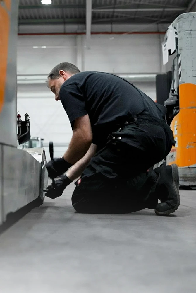 A service technician in black workwear kneels on the floor of a warehouse and works on an orange and grey industrial truck with a tool.