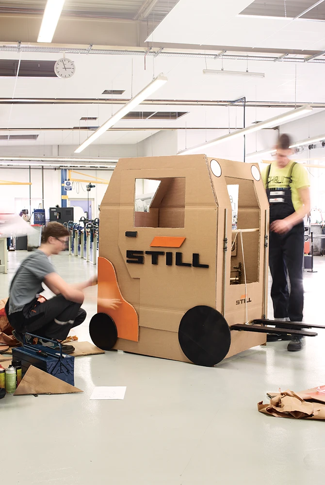 Two young people build a life-size forklift out of cardboard with the STILL logo in a workshop.