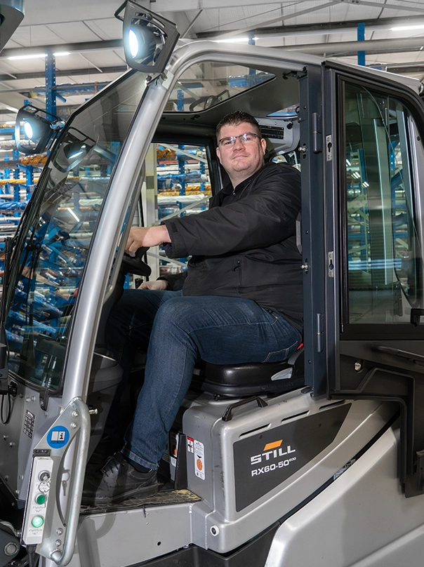 A man with glasses sits in the cab of a STILL RX60-50 forklift with the door open, in a warehouse with blue racks.