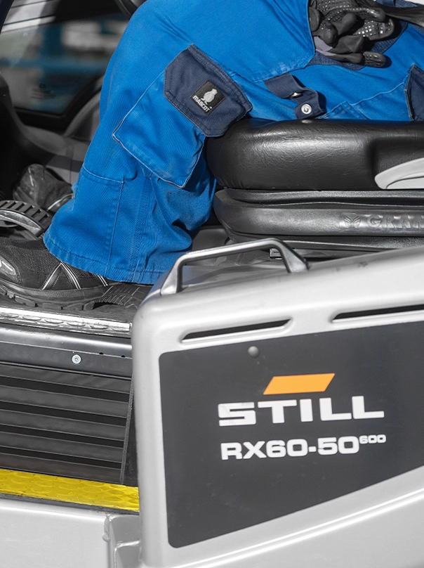 A driver in blue workwear sits on the seat of a STILL RX60-50 forklift. Only the legs and the side of the truck with the model name are visible.