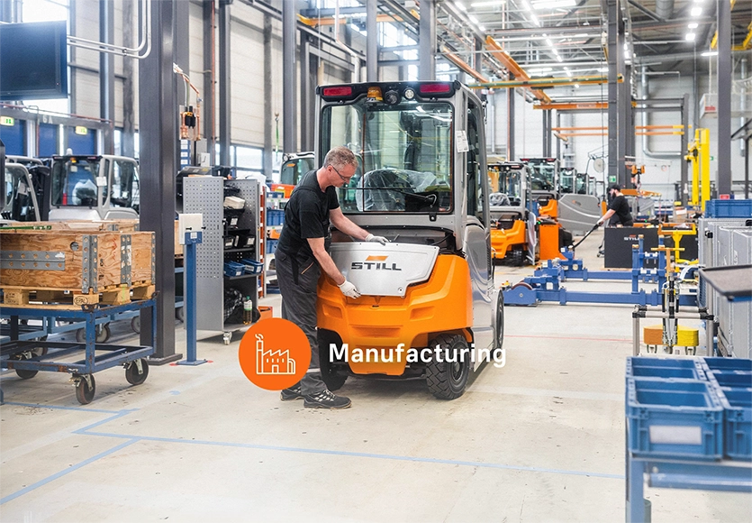A worker fits a part to the rear of an orange and grey STILL forklift in a production hall, labelled “Manufacturing".