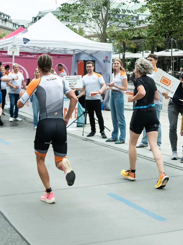 Two people run past a STILL tent, where people in STILL shirts cheer them on with signs.
