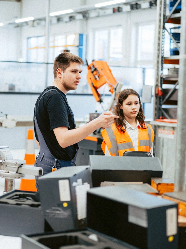 A young man in work overalls explains something in a production hall to a girl in an orange high-visibility vest.