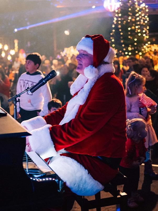 A man dressed as Santa Claus plays the piano and sings at a Christmas event, with children and a lit Christmas tree in the background.