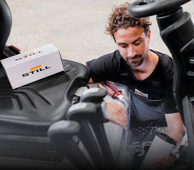 A STILL service technician with a filter cartridge in his hand works in the engine compartment of a forklift, with a STILL spare parts box on the seat.