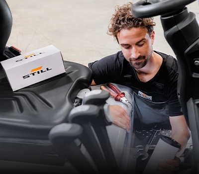 A STILL service technician with a filter cartridge in his hand works in the engine compartment of a forklift, with a STILL spare parts box on the seat.